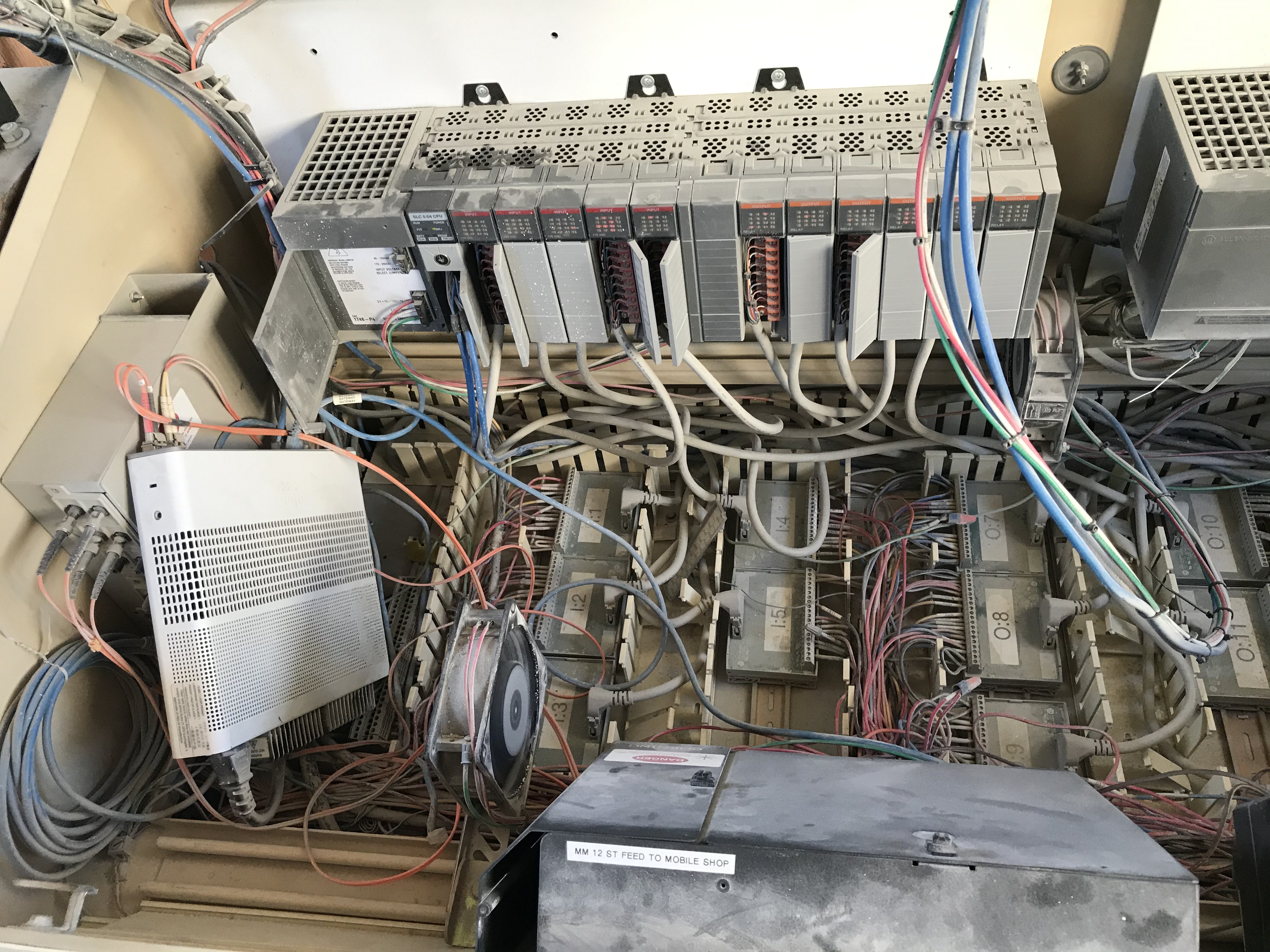 Dust-covered legacy PLC rack above relays, cable bundles, and cooling fans inside a control cabinet.